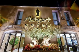 L'esterno dell'Hotel Melqart, con un cartello ben visibile, un albero imponente e un'aiuola fiorita di fronte.