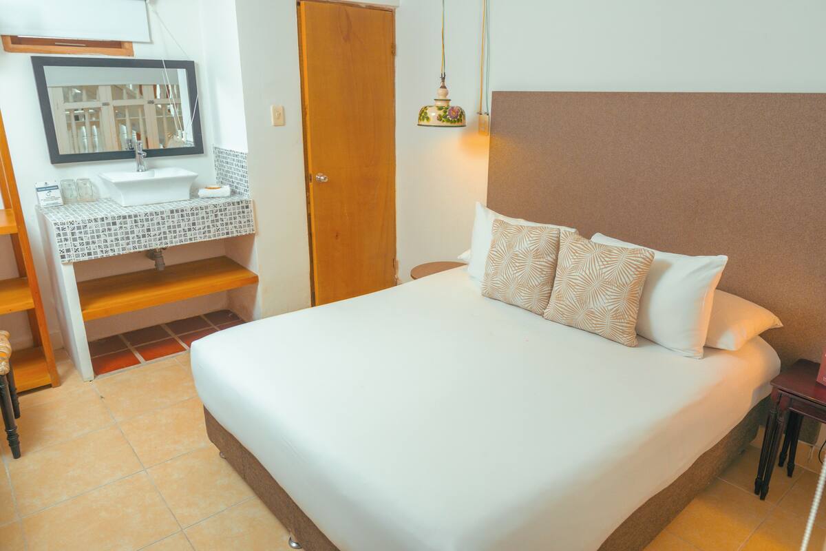 A bedroom with a large bed, a bedside table, a nightstand, a wooden door, a bathroom with a sink and mirror, and a hanging light fixture.