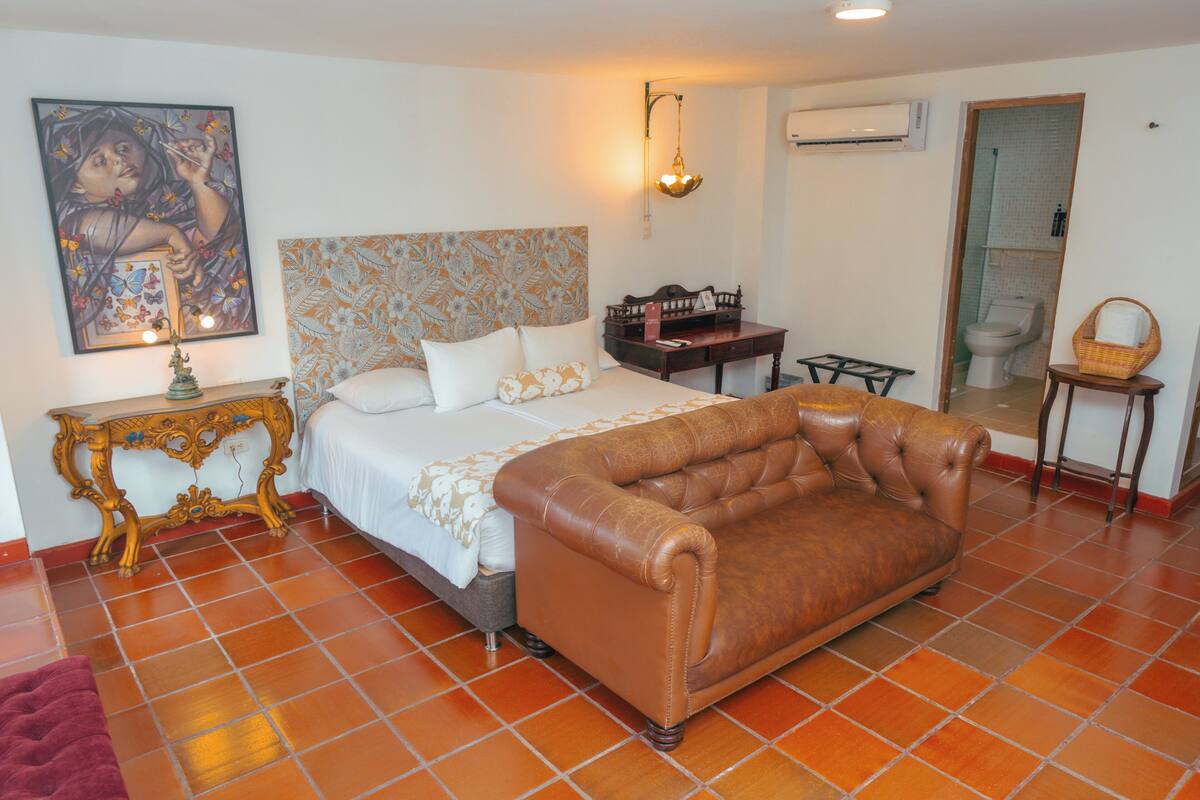 A bedroom with a bed, a brown leather sofa, a nightstand, a wall-mounted lamp, and a painting.