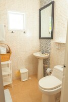 A small bathroom with a toilet, sink, and a mirror. There is a basket on a shelf and a small trash bin.