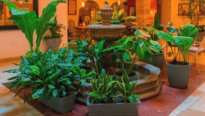 Un espace intérieur vaste et lumineux, agrémenté de nombreuses plantes en pot, d’une fontaine en pierre et d’un balcon doté de balustrades blanches.