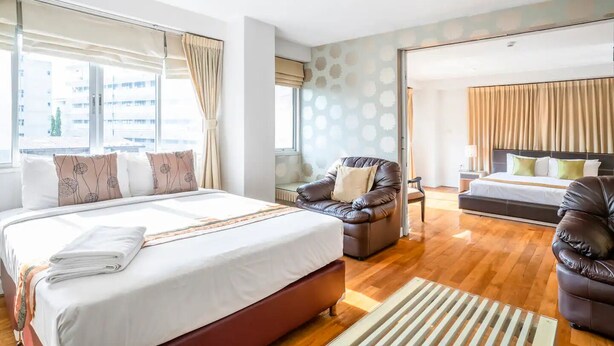 Quarto de hotel moderno com uma cama grande, uma poltrona de couro, um sofá e vista da cidade através de amplas janelas.
