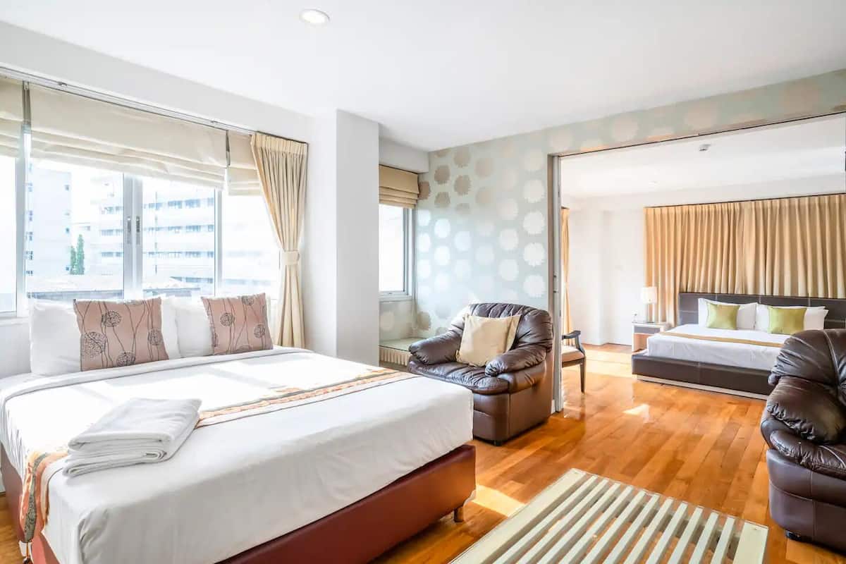 A modern hotel room with a large bed, a leather armchair, a sofa, and a view of the city through large windows.