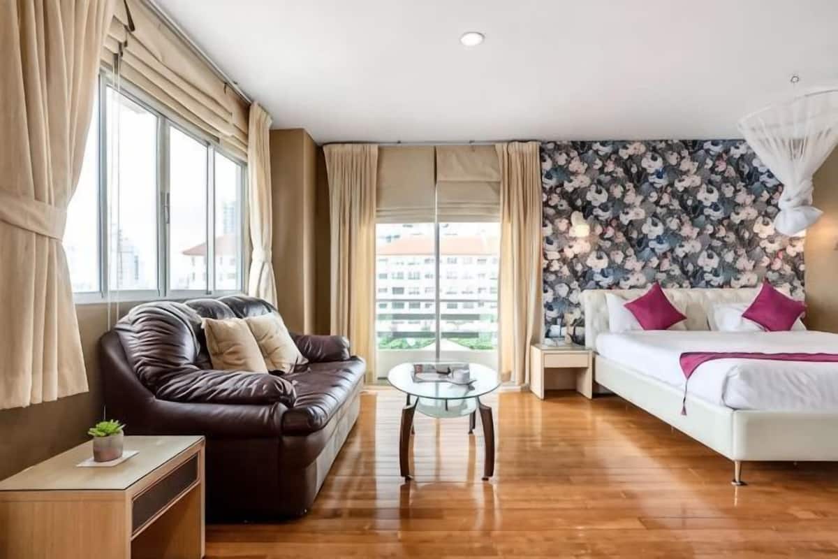 A room with a sofa, a bed, a glass coffee table, and a view of buildings outside.