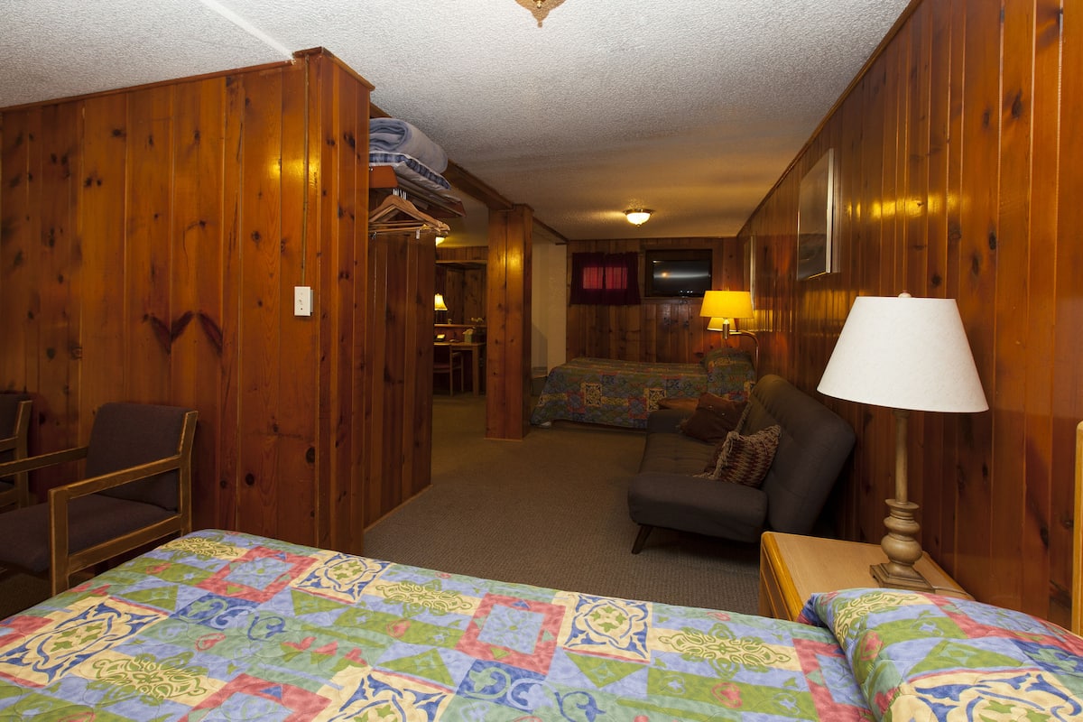 A hotel room with wooden walls, a bed with a colorful quilt, a sofa, a chair, and a lamp.