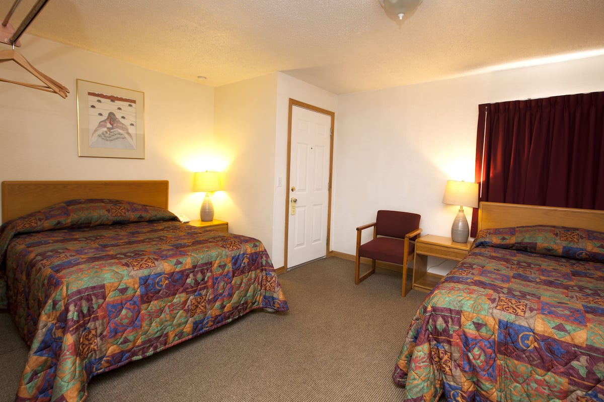 A hotel room with two beds, a chair, a desk with a lamp, and a door.