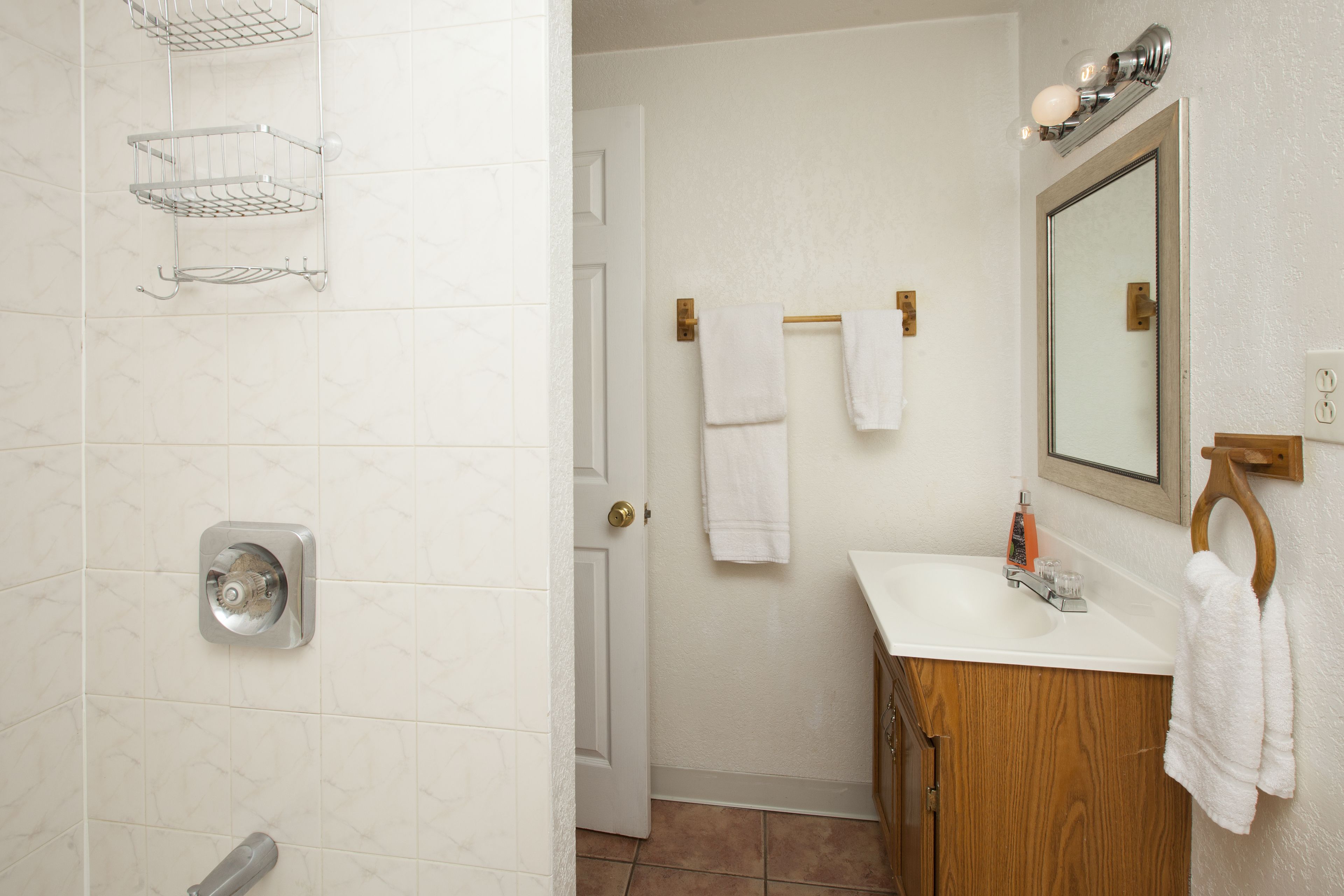 A bathroom with a white tiled wall, a wooden vanity, a mirror, a towel rack, and a shower with a chrome showerhead.