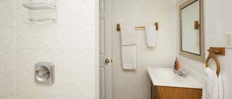 A bathroom with a white tiled wall, a wooden vanity, a mirror, a towel rack, and a shower with a chrome showerhead.