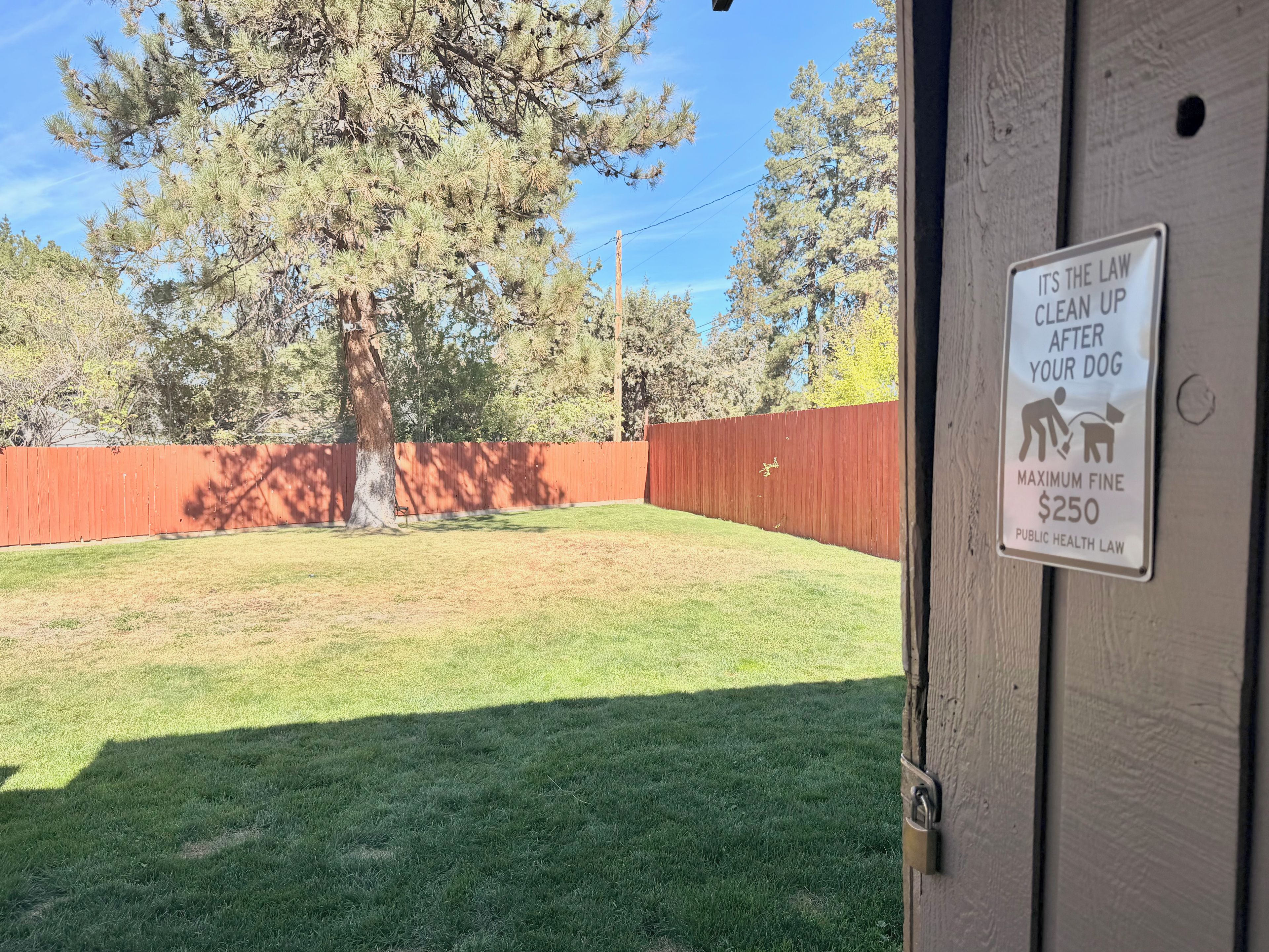 A backyard with a wooden fence, a tree, and a sign on a door warning about dog waste fines.