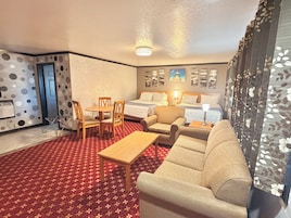 A hotel room with a bed, a sofa, a dining table with chairs, a small table with a lamp, a wall with a picture, and a patterned carpet.