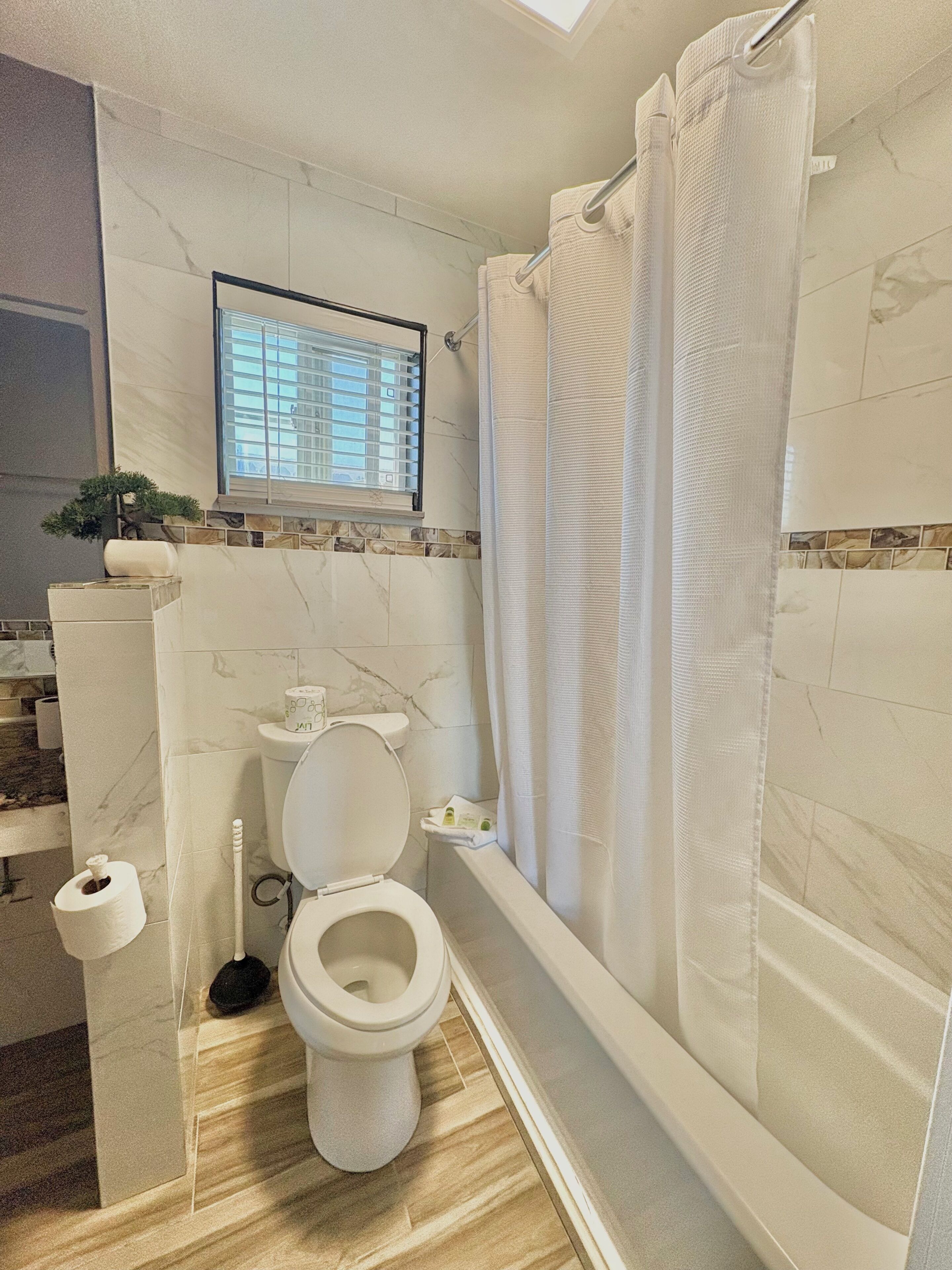 A bathroom with a toilet, a shower with a curtain, a window with blinds, and a small plant.