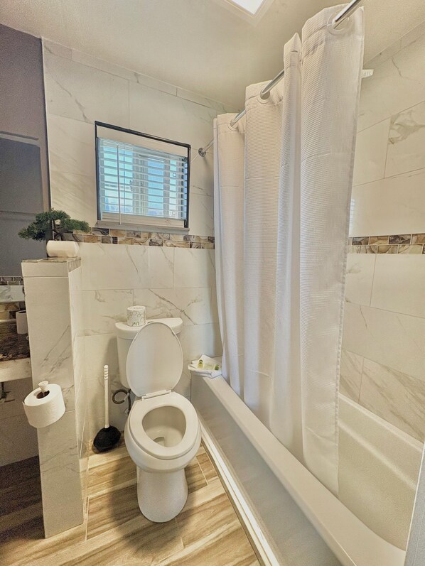 A bathroom with a toilet, a shower with a curtain, a window with blinds, and a small plant. - Cascade Lodge Boutique (Bend)