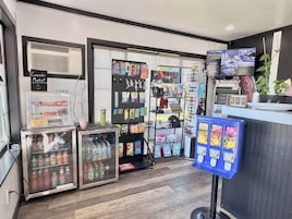 A small convenience store with a refrigerated drink cooler, a vending machine, and a wall-mounted security camera system.