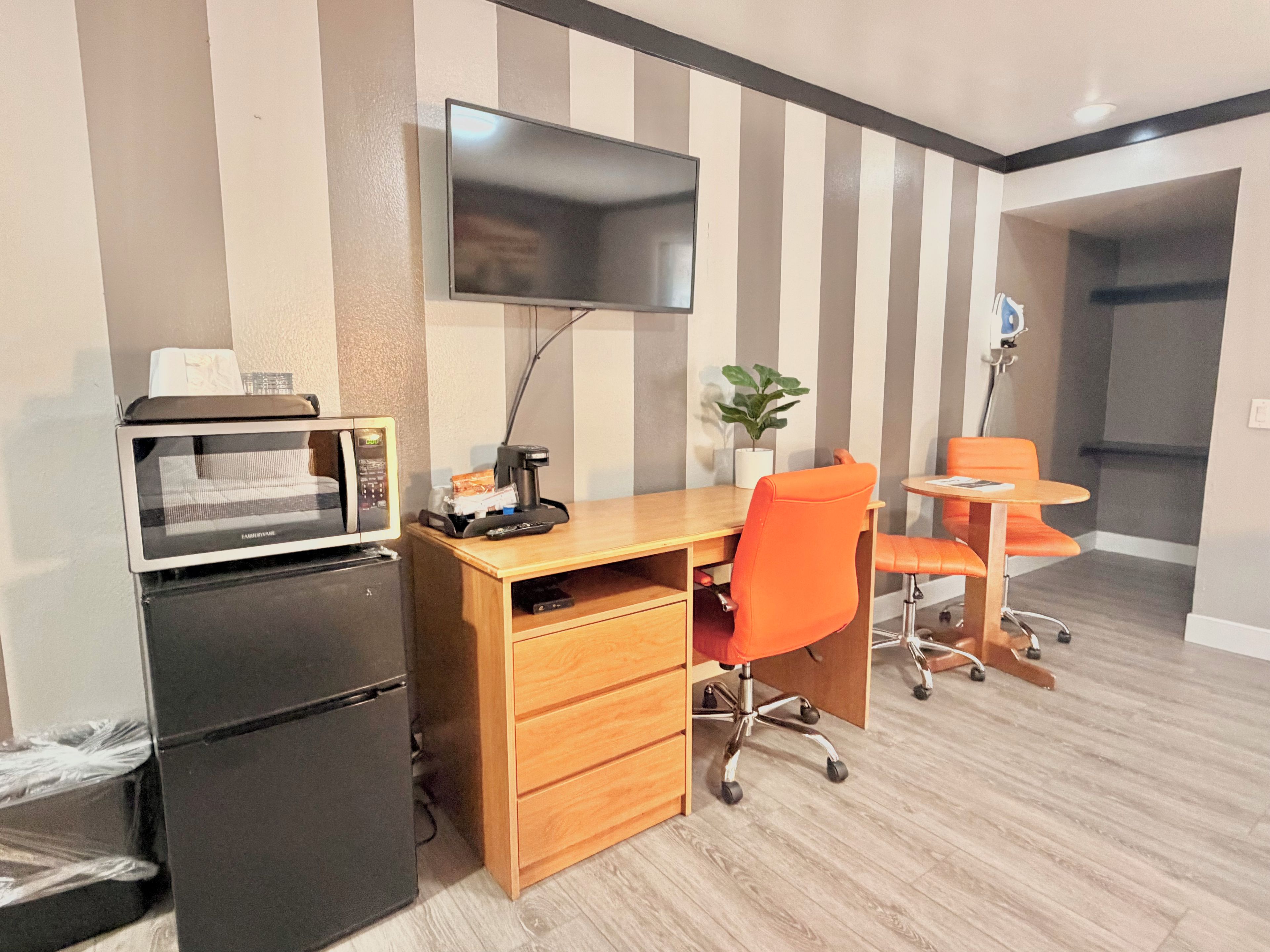 Een compacte hotelkamer met kitchenette, een flatscreen televisie aan de muur, een houten bureau met oranje bureaustoel en een kleine eettafel met passende stoelen.