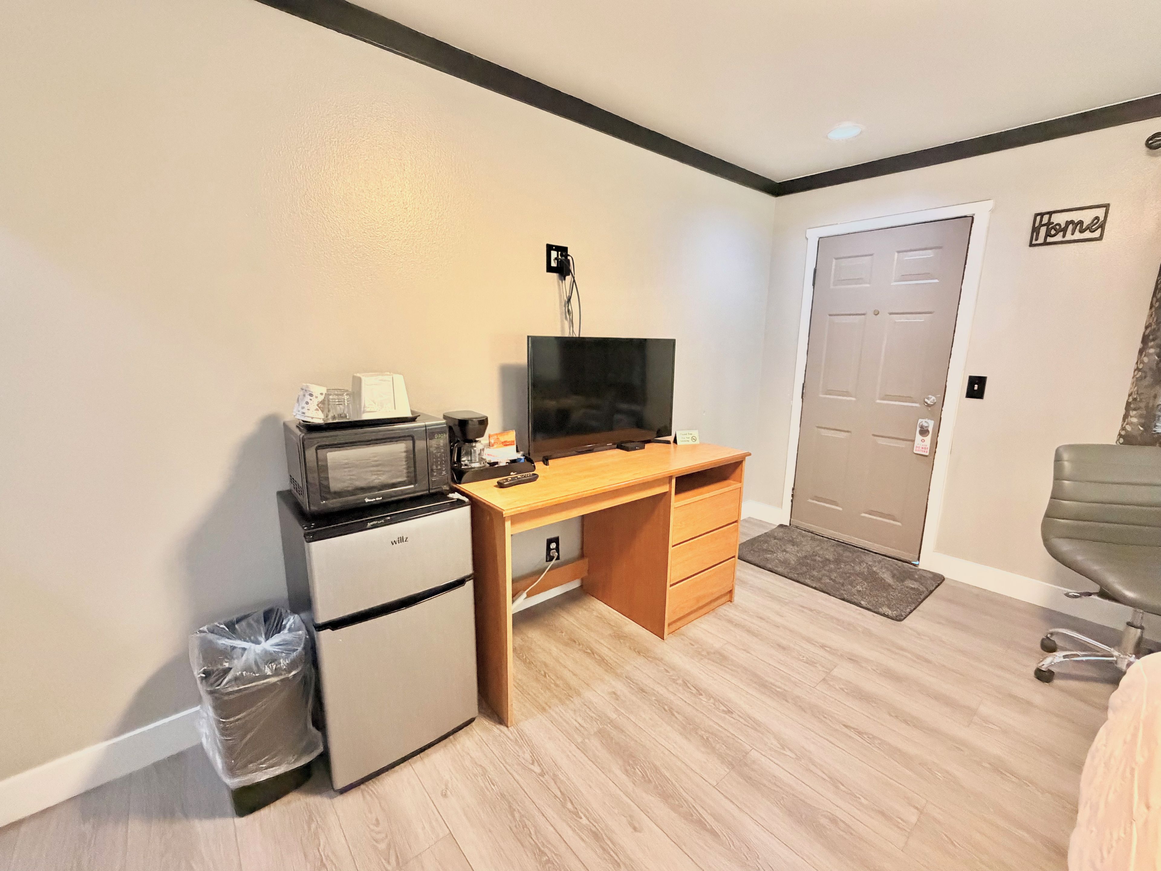 A compact room with a kitchenette, a wooden desk, a television, and a door labeled 'Home'.