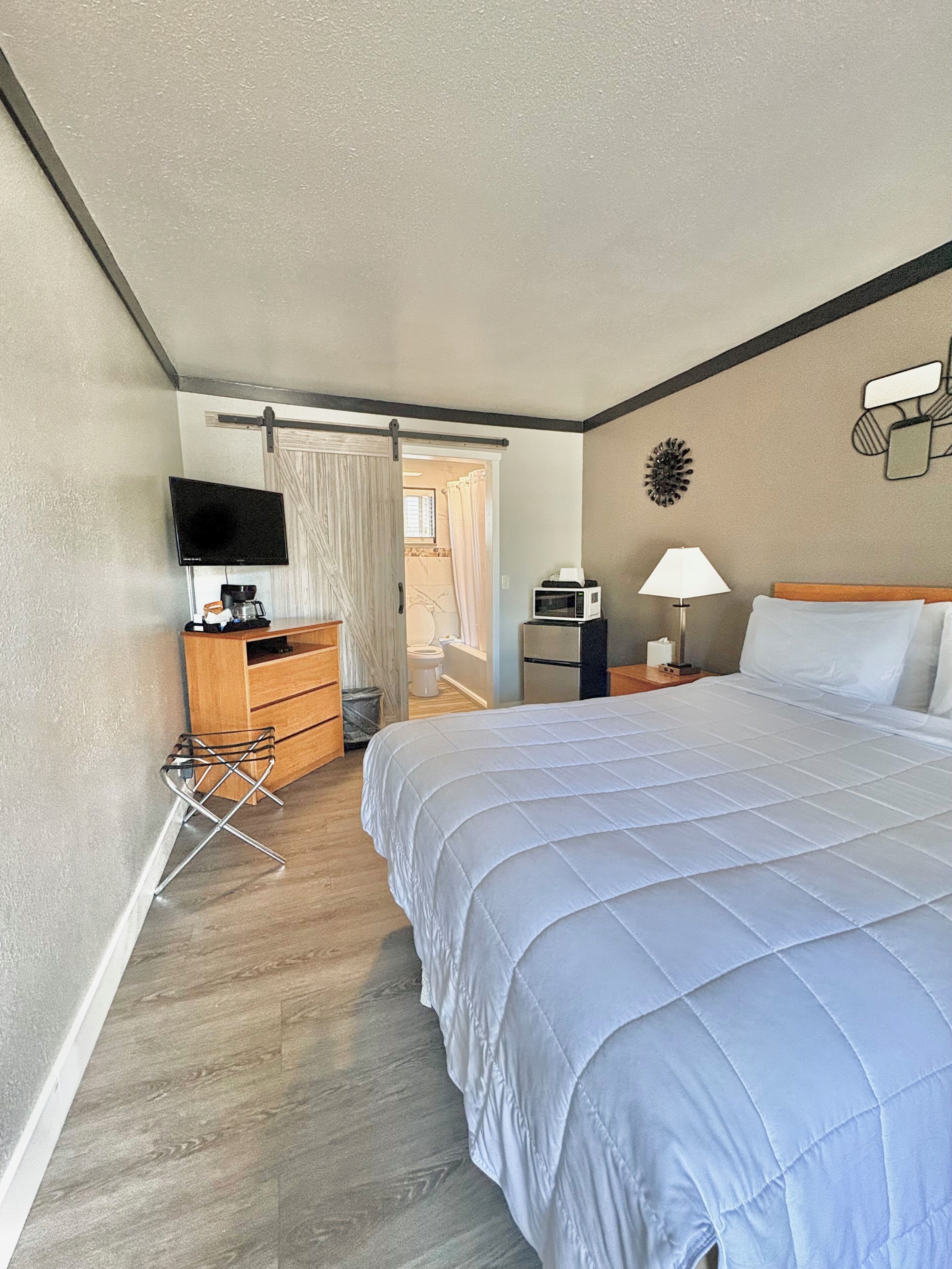 A hotel room with a bed, a desk with a computer, a television, and a bathroom visible through an open door.