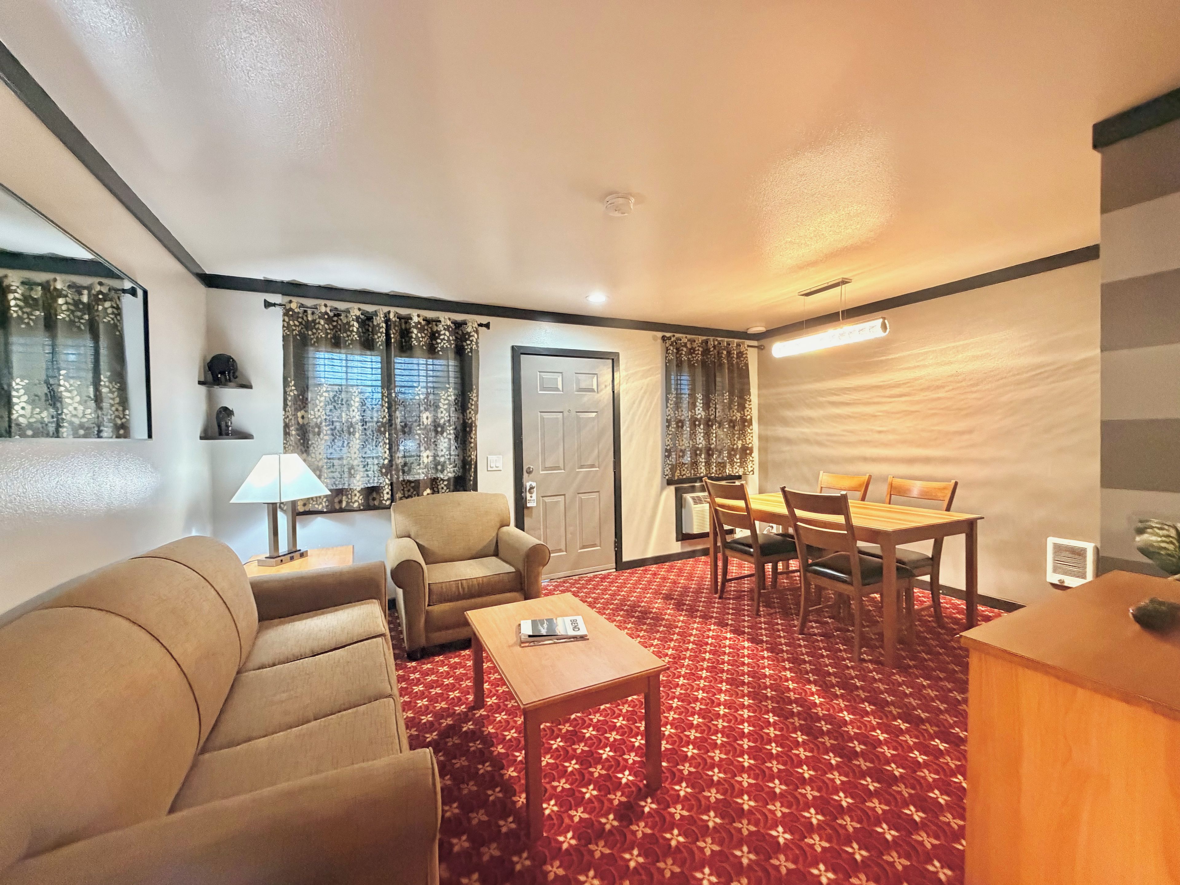 A hotel room with a sofa, armchairs, a dining table with chairs, a lamp, and a patterned carpet.