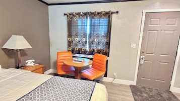 A hotel room with a bed, a desk with a lamp, a telephone, a small round table with two orange chairs, and a window with floral curtains.