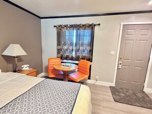 A hotel room with a bed, a desk with a lamp, a telephone, a small round table with two orange chairs, and a window with floral curtains. - Cascade Lodge Boutique (Bend)