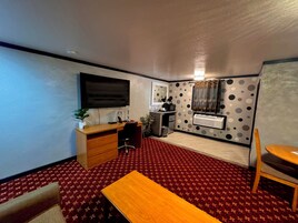 Habitación compacta con escritorio de madera, una silla, un televisor montado en la pared, una kitchenette pequeña y una alfombra estampada.
