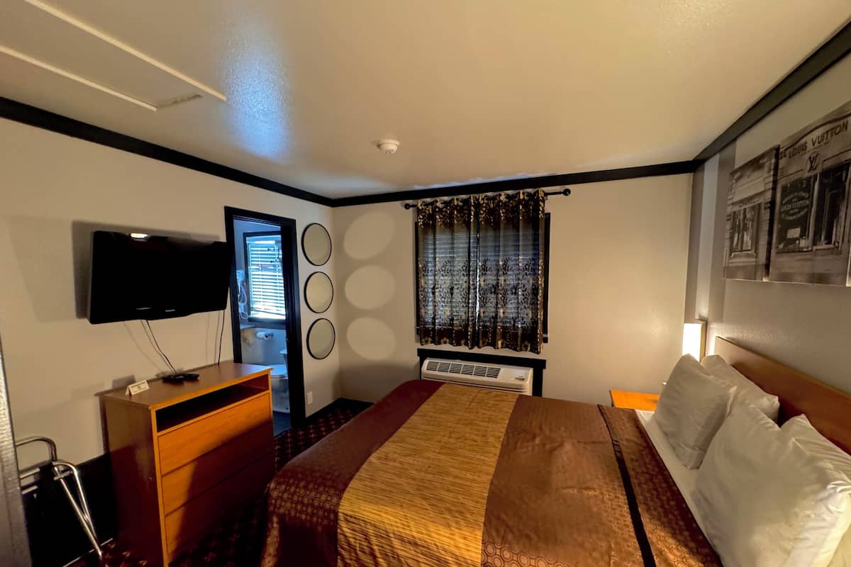 A hotel room with a bed, a TV mounted on the wall, a dresser, a window with curtains, and a wall-mounted air conditioner.