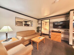 Habitación de hotel pequeña con sofá, una mesita, escritorio con televisor, espejo y puerta que da a un dormitorio.