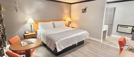 A hotel room with a bed, two bedside lamps, a round wooden table, an orange chair, and a small desk with a chair.