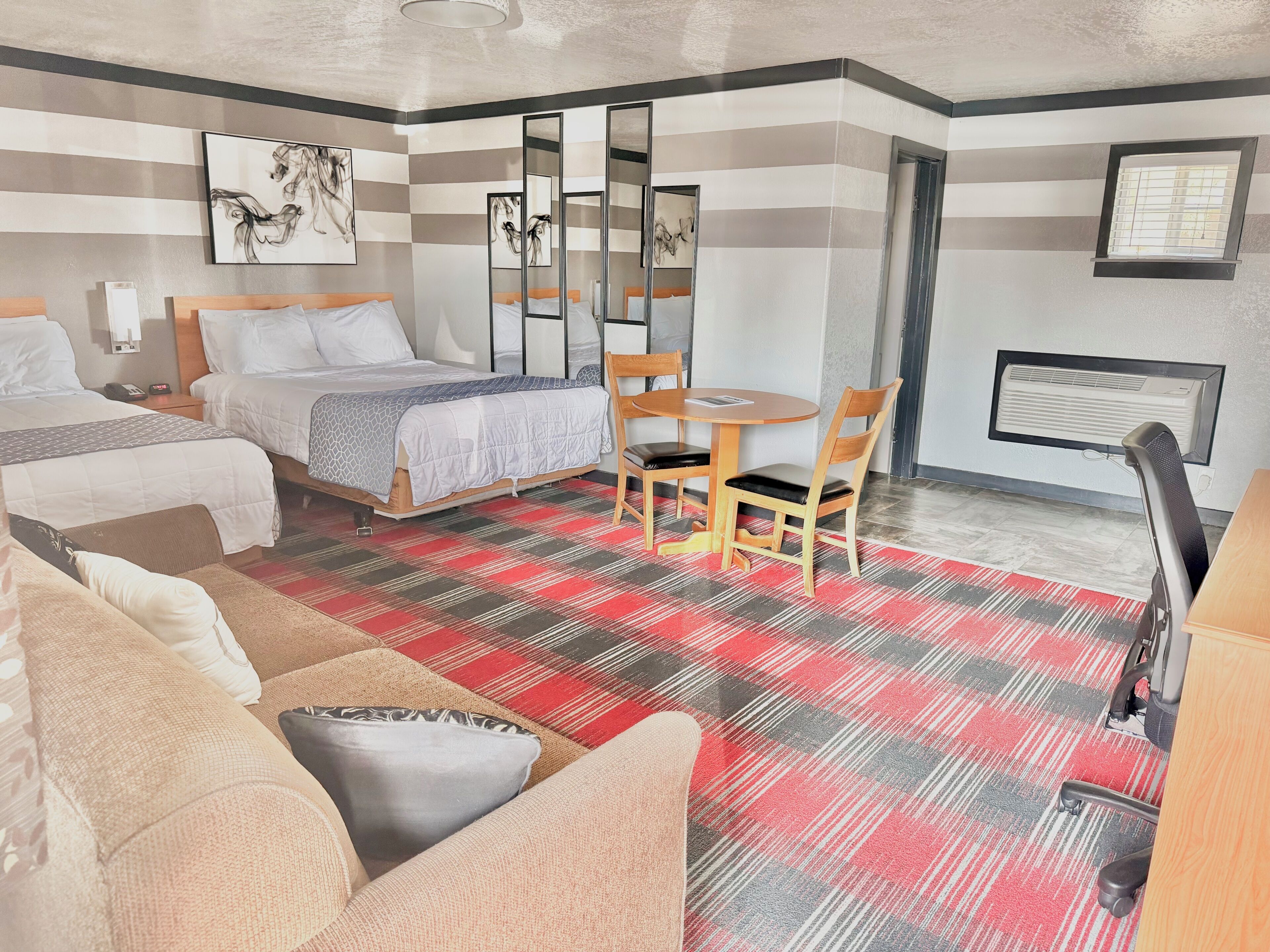 A hotel room with two beds, a dining table with chairs, a sofa, a mirror, artwork on the wall, and an air conditioning unit.