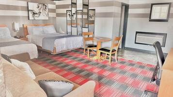 A hotel room with two beds, a dining table with chairs, a sofa, a mirror, artwork on the wall, and an air conditioning unit.