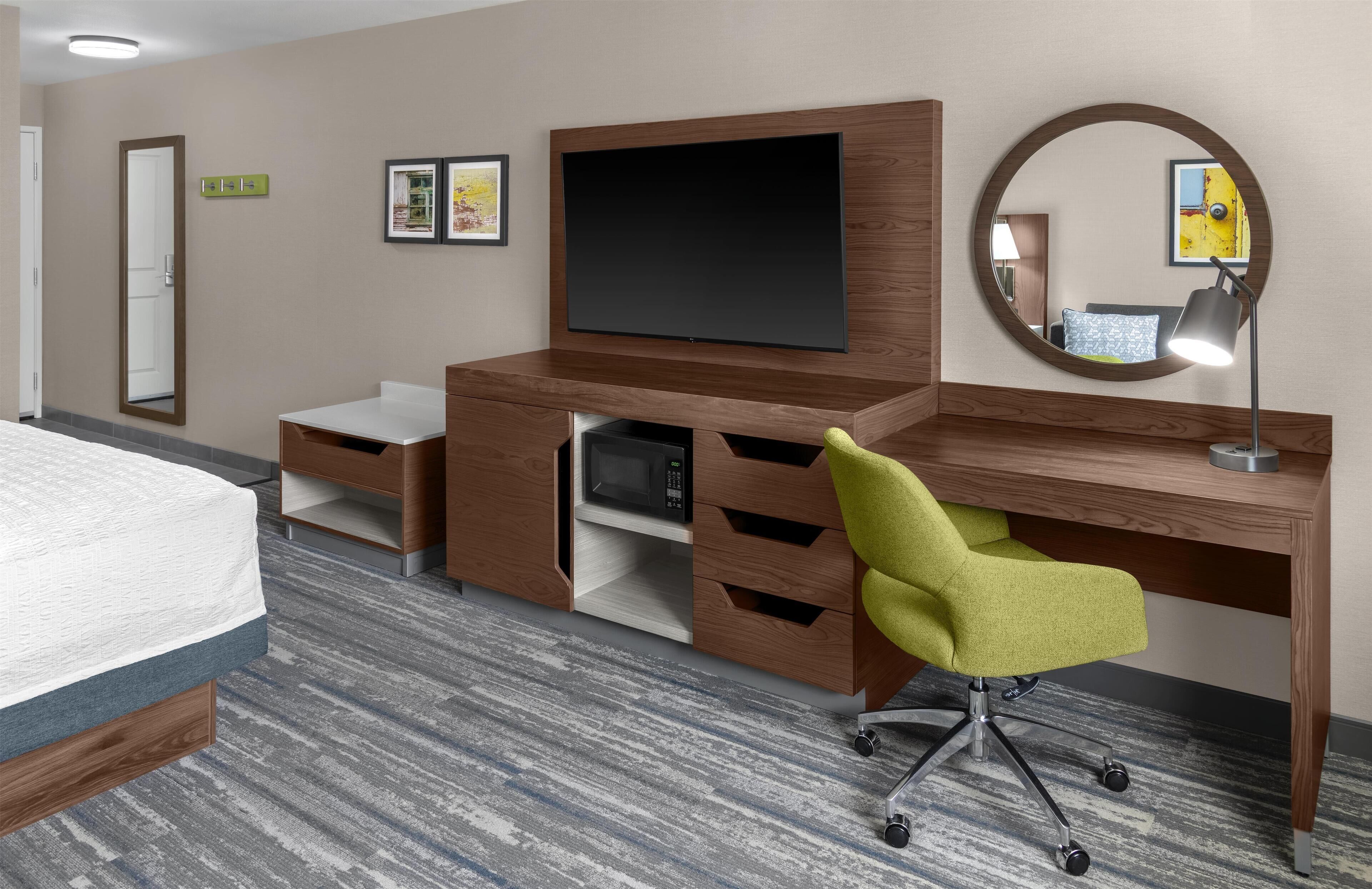 A hotel room with a bed, a desk with a chair, a flat-screen TV, a mirror, and a small shelf.