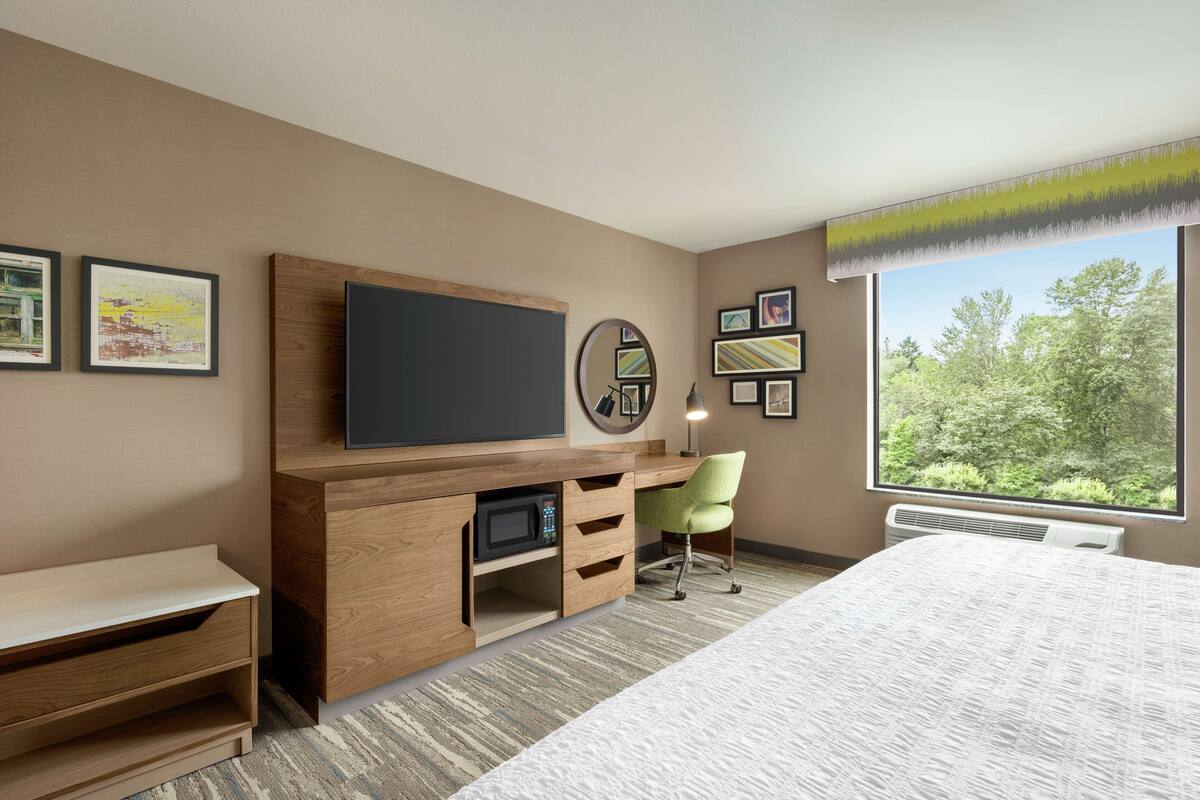 A hotel room with a bed, a desk with a flat-screen TV, a microwave, a chair, and a large window with a view of trees.