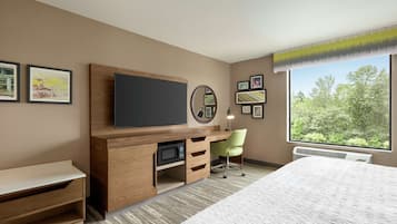 Habitación de hotel con cama, escritorio, televisor de pantalla plana, microondas, silla y ventana grande con vistas a árboles.