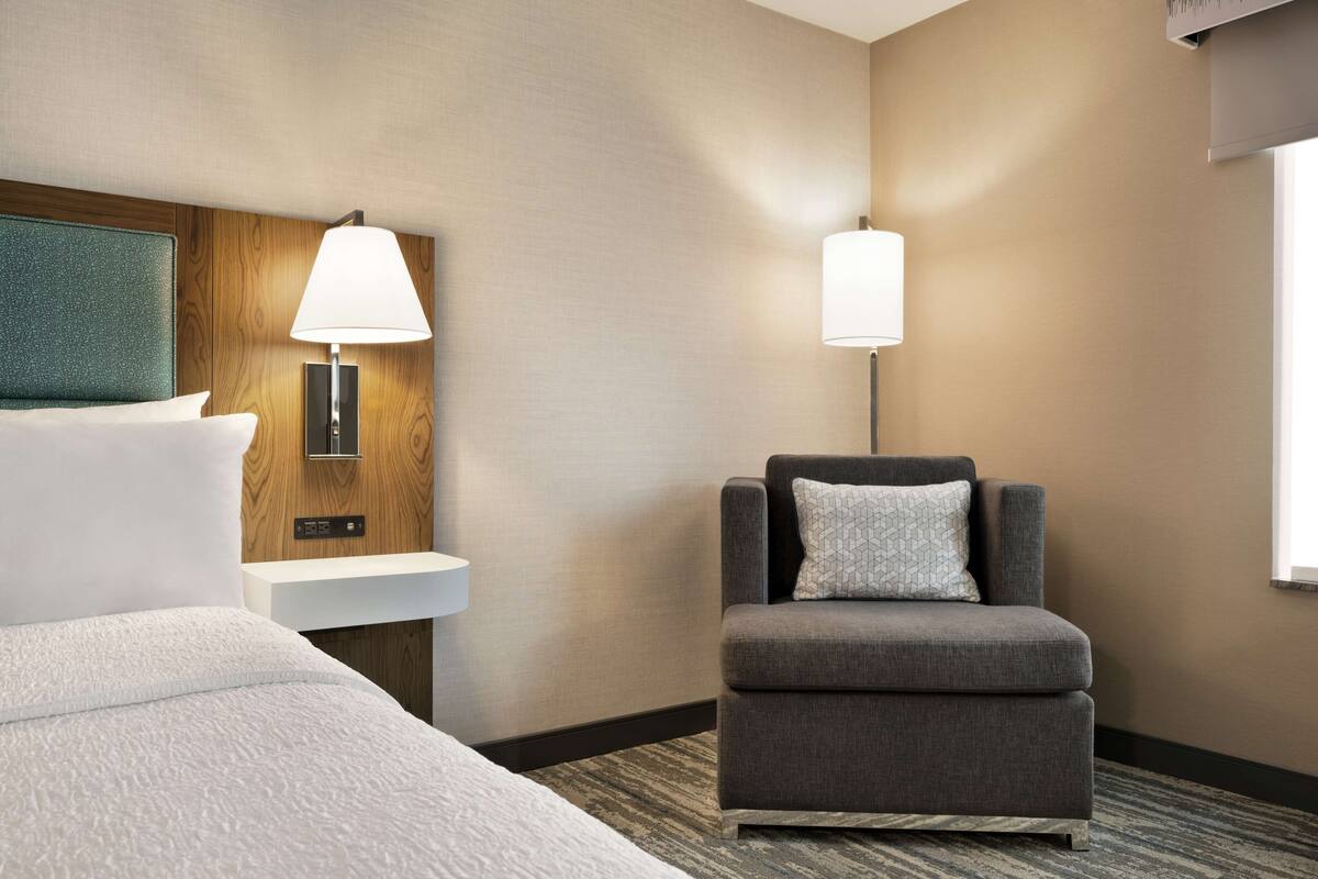 A hotel room with a bed, a grey armchair, a wooden headboard, a white bedside table, and two wall-mounted lamps.