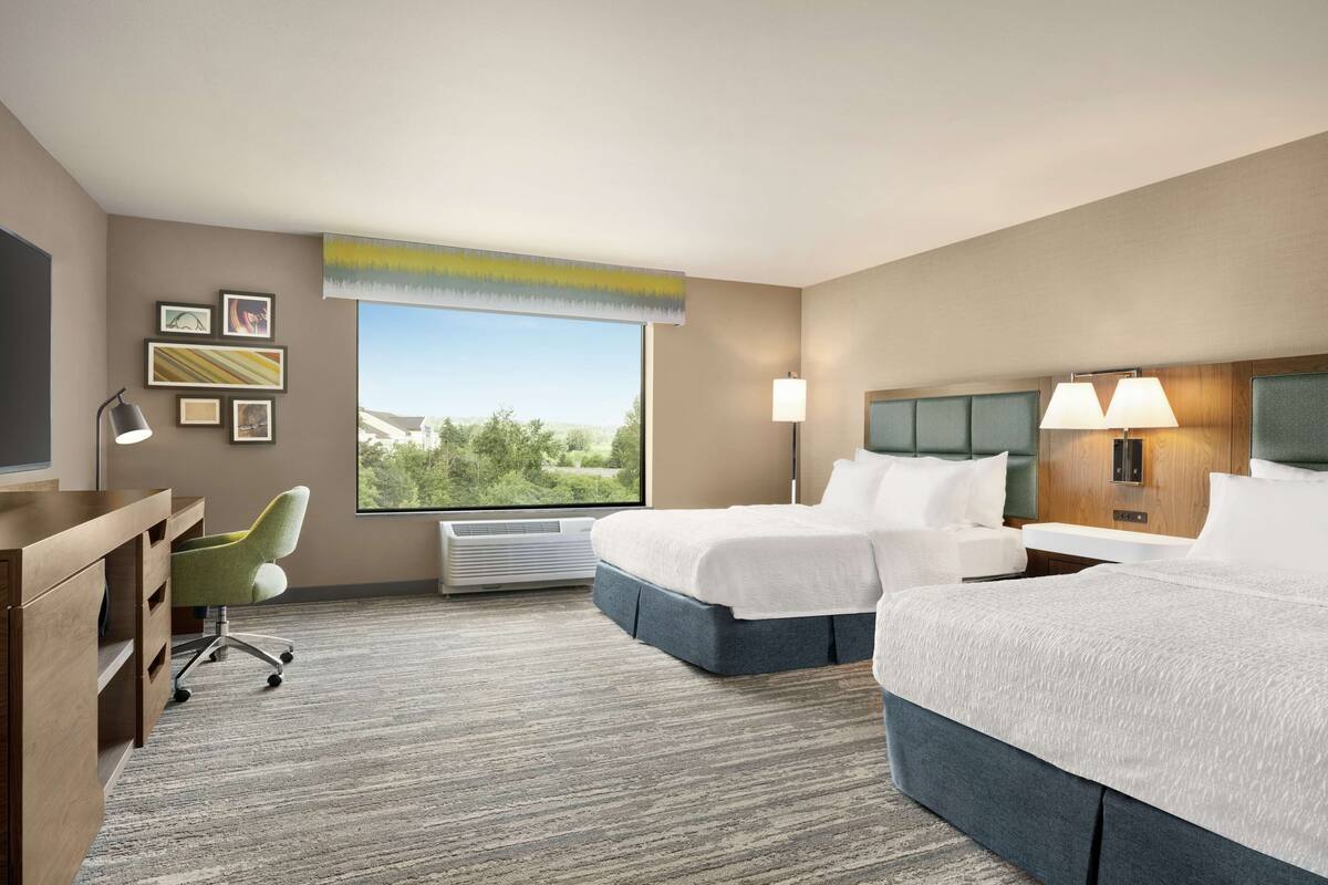 A hotel room with a large bed, a desk, a chair, and a window with a view of greenery.