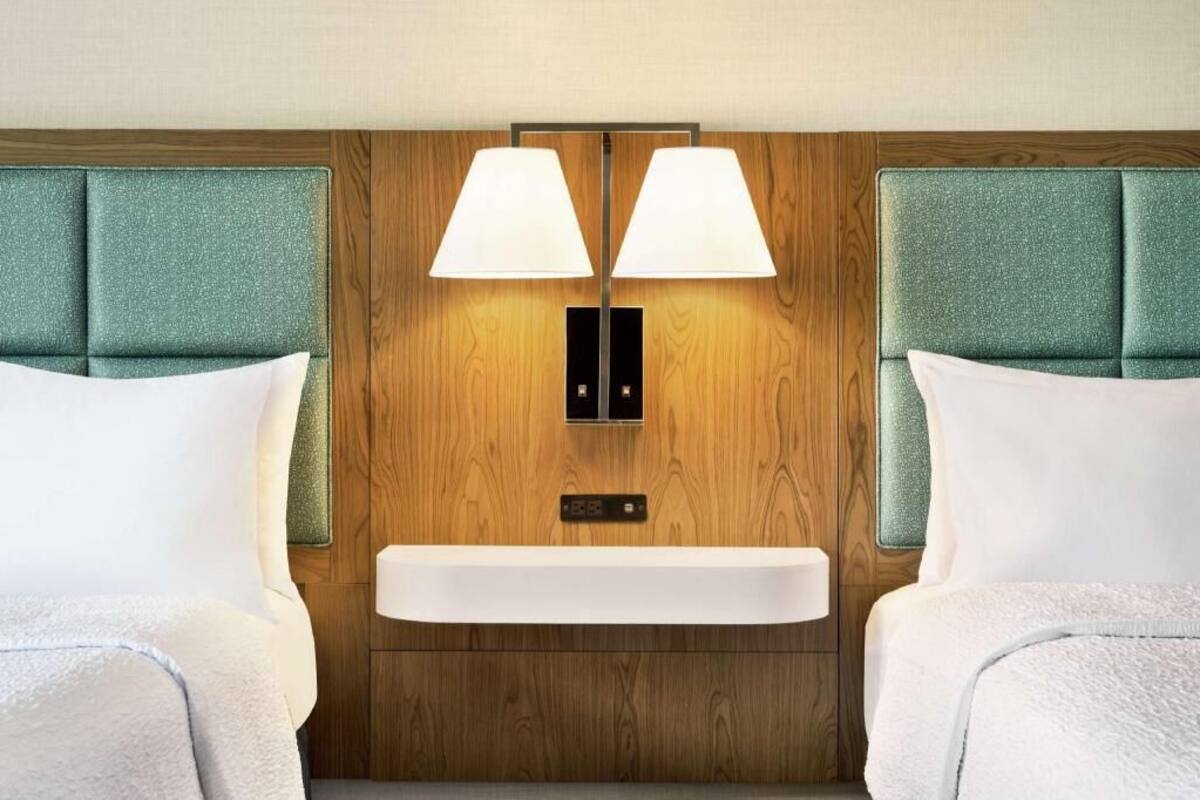 A hotel room with a wooden headboard, two bedside lamps, a built-in shelf, and a bed with white bedding.