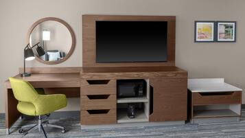 A modern hotel room with a wooden desk, a green office chair, a flat-screen TV, a microwave, a phone, and a small drawer unit.