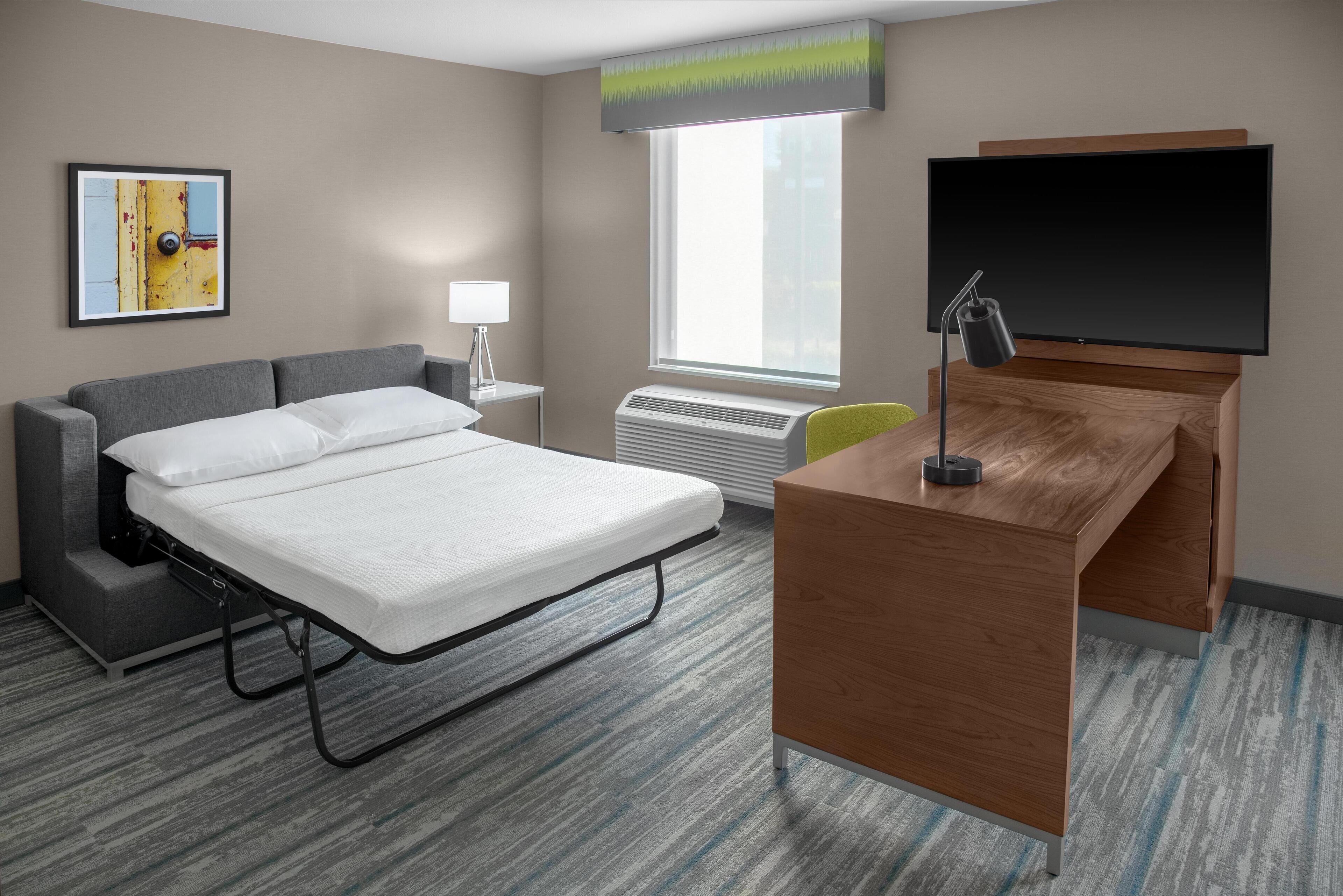 A hotel room with a bed, a desk, a chair, a TV, a window, and a painting on the wall.