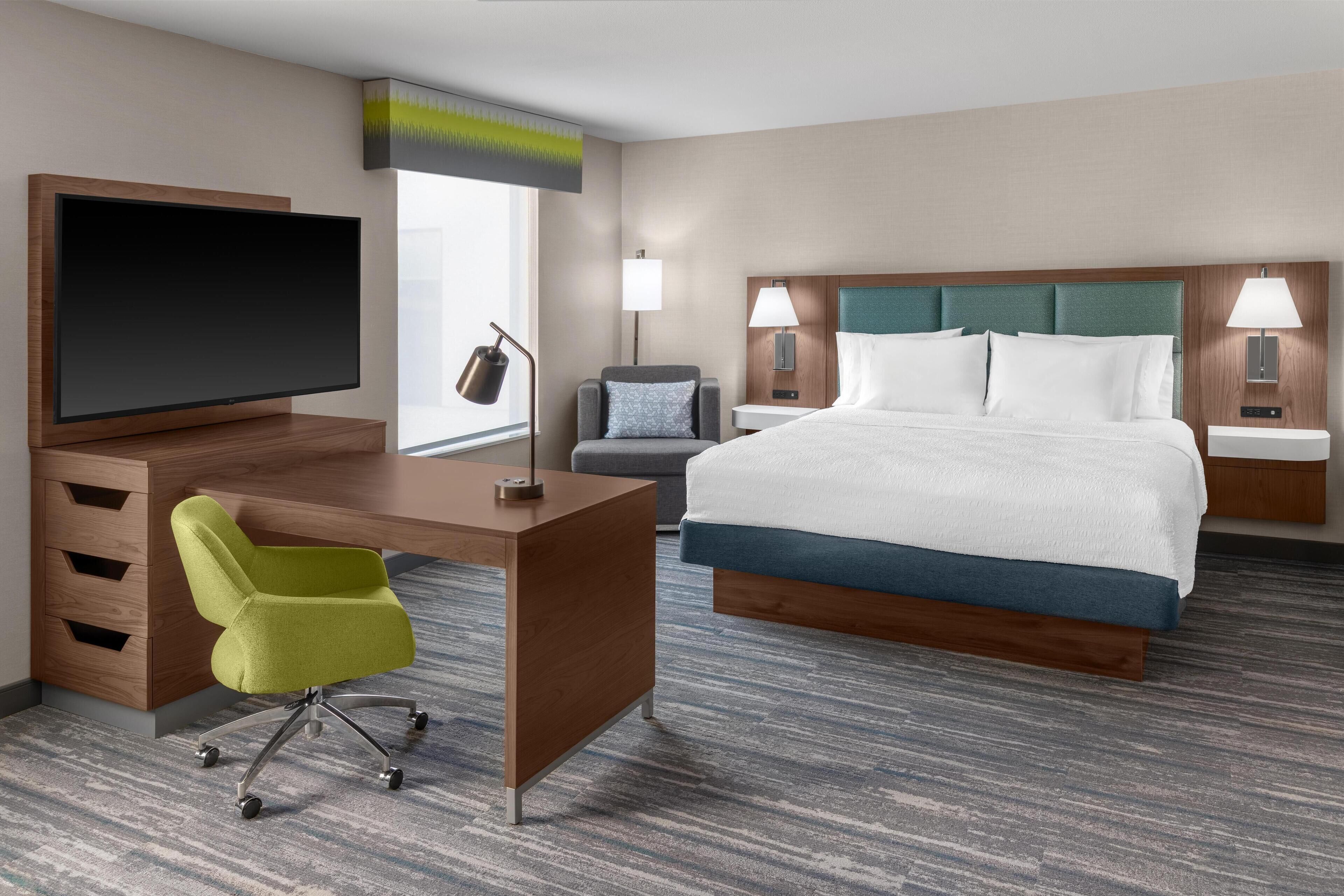 Habitación de hotel con cama, escritorio, silla, televisor, lámparas y ventana.