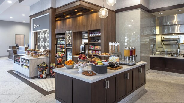 Eine moderne Hotelhalle mit einem Selbstbedienungs-Frühstücksbereich, einer Rezeption und einem gut sichtbaren Schild mit der Aufschrift 'The Shop'.