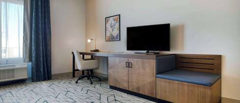 Habitación de hotel con escritorio, televisión, banca acolchada y alfombra estampada.