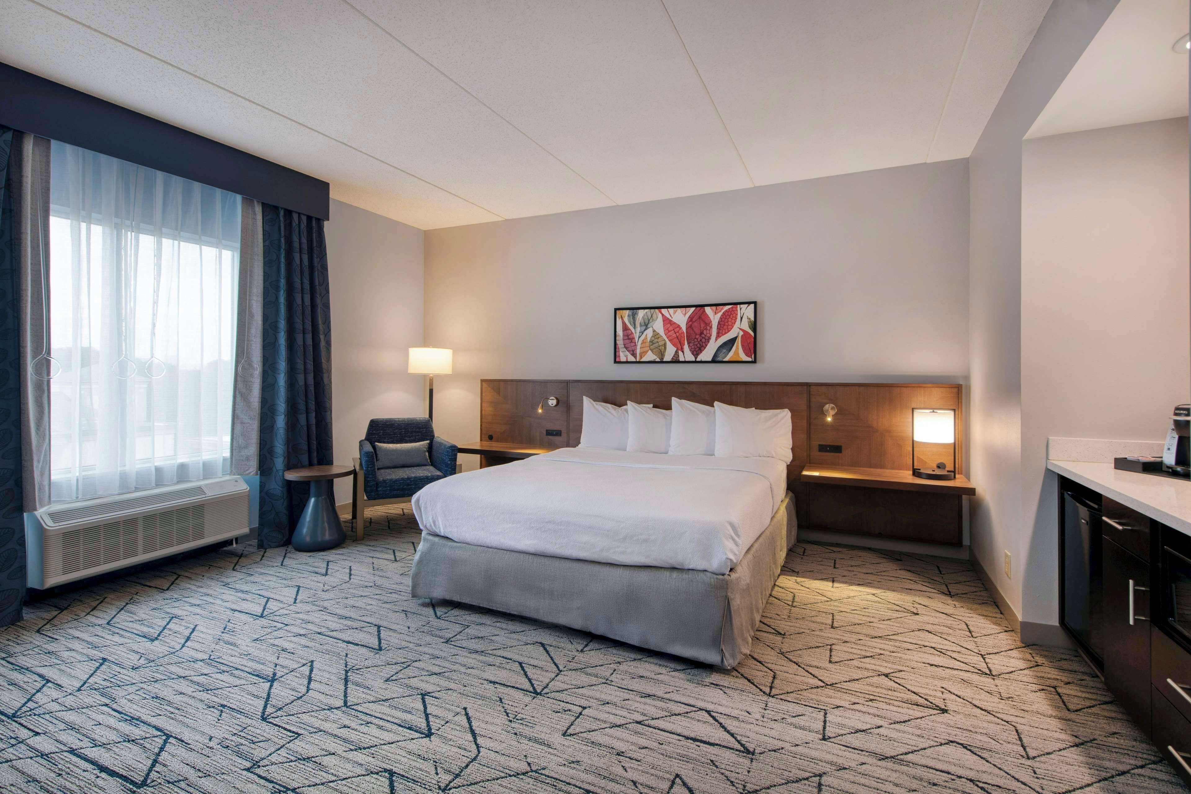 Une chambre d’hôtel avec un grand lit, deux tables de chevet avec des lampes, une chaise, un bureau avec une chaise, un radiateur et une œuvre d’art accrochée au mur.