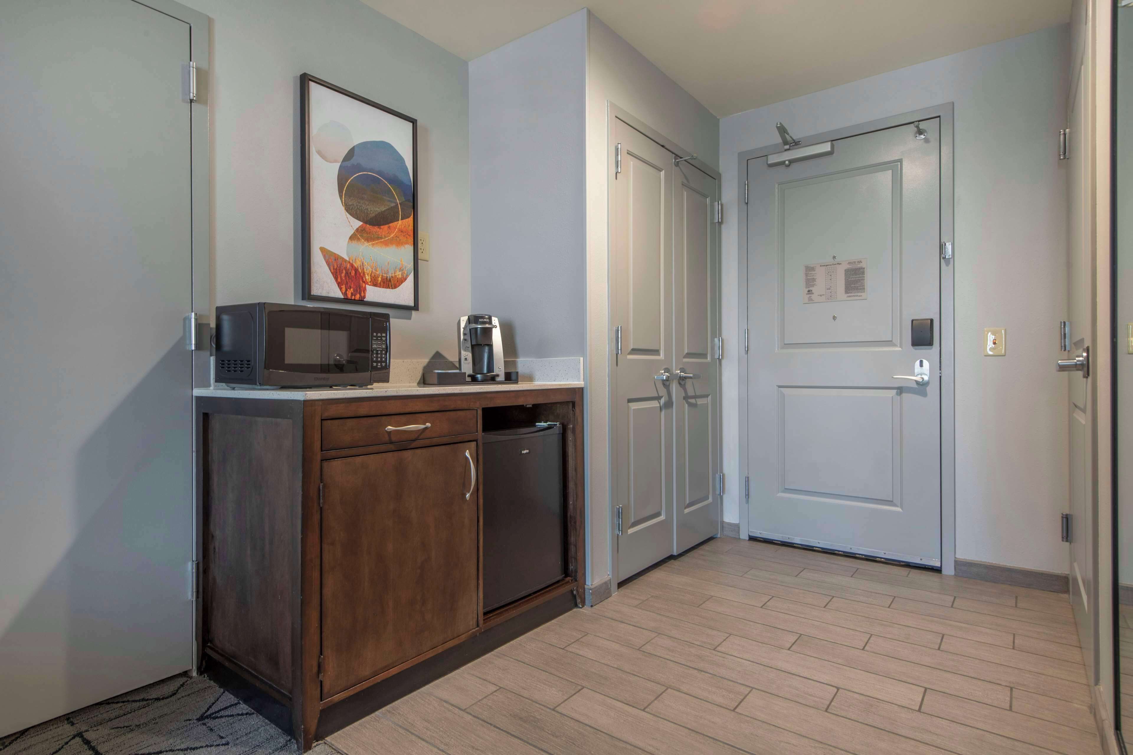 A compact kitchen with a microwave, coffee maker, and a refrigerator. There are two doors, one with a calendar and the other with a light switch. The floor is wooden.