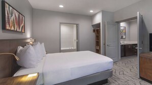 A modern hotel room with a large bed, a bedside table, a desk, a bathroom with a sink and mirror, and a walk-in closet.