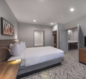 A modern hotel room with a large bed, a bedside table, a desk, a bathroom with a sink and mirror, and a walk-in closet.