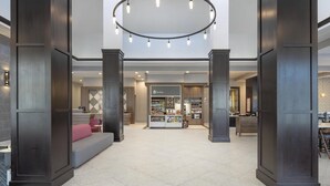 Eine großzügige Lobby mit einer zentralen Sitzgruppe, einem Empfangsbereich und einem Geschäft im Hintergrund mit dem Namen 'The Shop'.