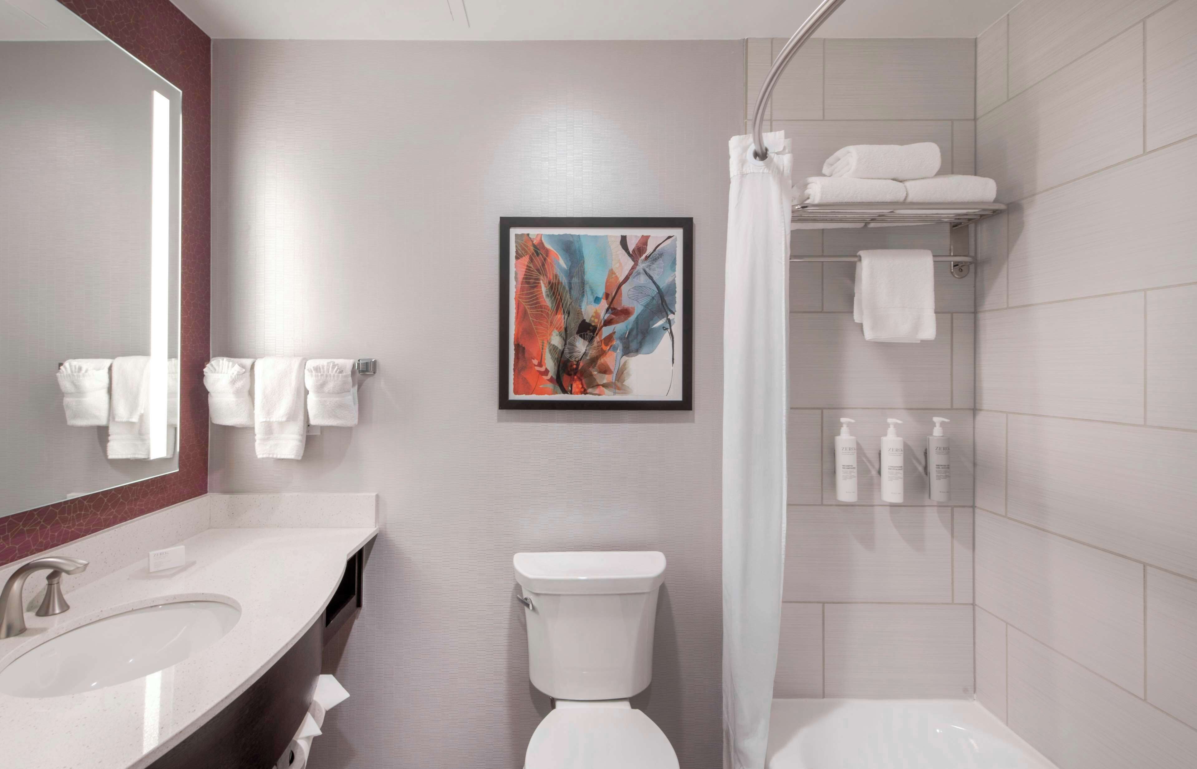 A modern bathroom with a sink, toilet, and shower. There is a framed artwork on the wall and a shelf with toiletries.