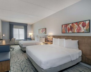 A hotel room with two beds, a desk, a chair, a window with curtains, and artwork on the walls.