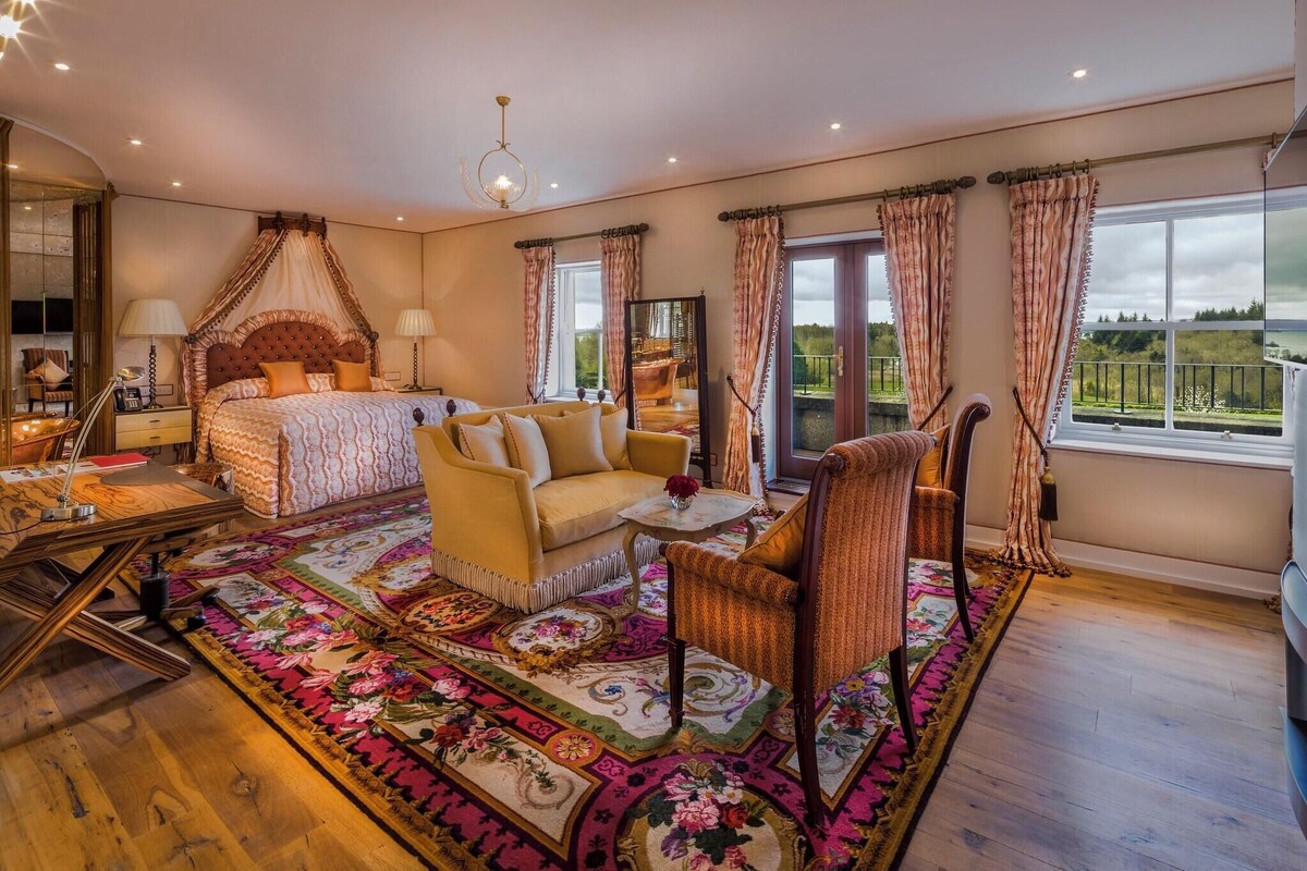 Ein Schlafzimmer mit einem Himmelbett, einer Couch, einem Sessel, einem kleinen Tisch und einem großen Fenster mit Vorhängen.