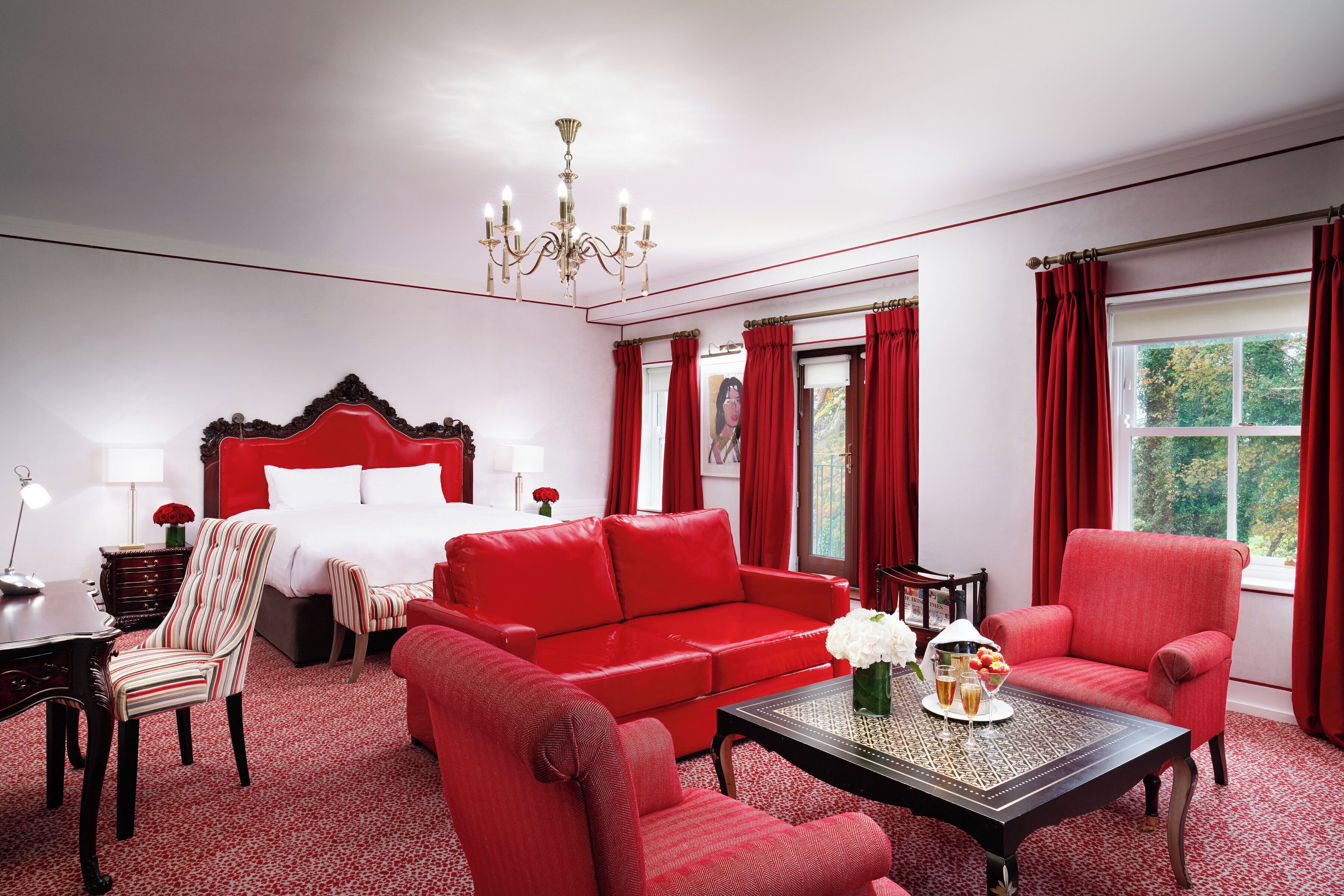 A hotel room with a large bed, red upholstered furniture, a chandelier, and a view of trees through the window.