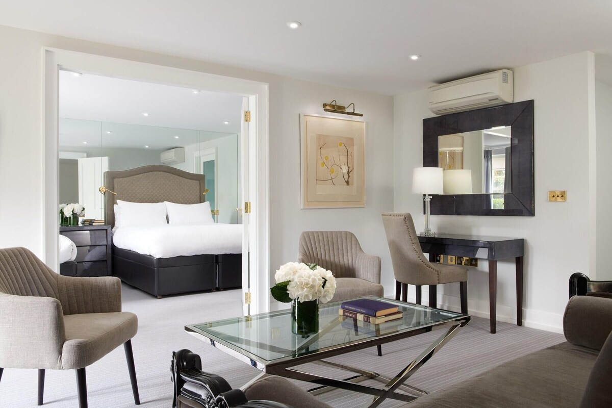Ein modernes Hotelzimmer mit einem Bett, einem Glastisch, zwei Sesseln, einem Schreibtisch mit Lampe und einem großen Spiegel.